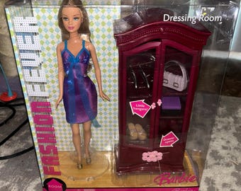 May include: A Barbie Fashion Fever doll in a blue and purple dress stands next to a red dressing room. The dressing room has a white purse, shoes, and a purple box inside. The packaging reads "Dressing Room".