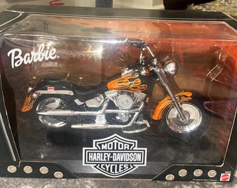 Pode incluir: Um brinquedo de motocicleta Harley-Davidson preto e laranja em uma vitrine de plástico transparente. A motocicleta tem chamas pintadas na lateral e as palavras "Harley-Davidson" e "Motor Cycles" estão impressas na parte inferior da vitrine.