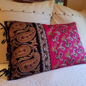 May include: A decorative pillow with a paisley pattern in black, gold, and brown on one side and a pink paisley pattern on the other. The pillow has black ties on the side and is placed on a white bed.