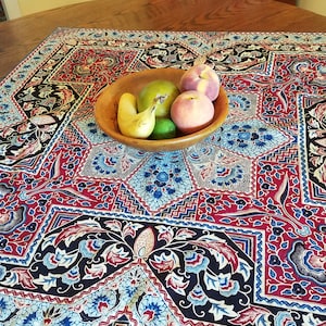 May include: A wooden bowl filled with fruit sits on a patterned tablecloth. The tablecloth features a red, blue, and black floral design. The fruit includes a pear, a lime, a peach, and a banana. The table is made of wood.