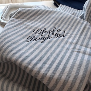May include: A white and grey striped kitchen towel featuring the embroidered black text Life of a Dough Girl. The fabric has a woven texture and is draped over an embroidery hoop, with a dark blue fabric visible in the background.