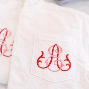 May include: White t-shirts with a pocket, each embroidered with a red and pink monogram. The monogram is a decorative script design. The shirts are folded, indicating a set or multiple items.