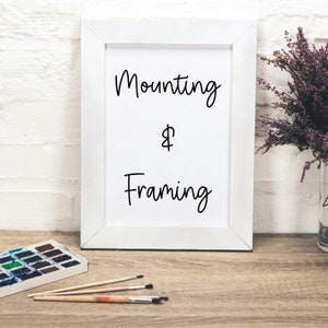 以下が含まれることがあります： 白いフレームに「Mounting & Framing」というテキストが書かれた白い背景。フレームは、水彩パレットと筆が置かれた木製の表面の上に置かれています。