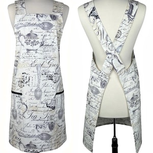 May include: A white apron with a vintage tea-themed design, featuring teapots, spoons, and script. The apron has black trim on the pockets and a cross-back design. The text "Tea Time" is visible on the front.