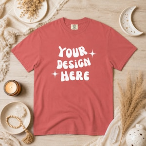 Puede incluir: Una camiseta de color coral con el texto blanco "YOUR DESIGN HERE" en letras estilizadas. La camiseta tiene cuello redondo y mangas cortas. El diseño incluye estrellas. La camiseta se muestra sobre una superficie de madera clara.