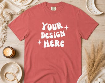以下が含まれることがあります： 白い「YOUR DESIGN HERE」の文字がスタイリッシュにデザインされた、コーラルカラーのTシャツ。Tシャツはクルーネックと半袖です。デザインには星のアクセントが含まれています。Tシャツは明るい木製の表面に表示されています。