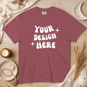 Puede incluir: Una camiseta granate con el texto "YOUR DESIGN HERE" en blanco, con letras estilizadas. Dos explosiones de estrellas blancas flanquean el texto. La camiseta se muestra sobre una superficie de madera con elementos decorativos.