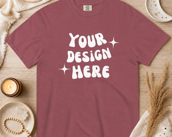 以下が含まれることがあります： 白の「YOUR DESIGN HERE」の文字がスタイリッシュにデザインされた、マルーン色のTシャツ。テキストの両側に白いスターバーストが配置されています。木製の表面に装飾的な要素とともにシャツが展示されています。