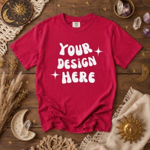 Puede incluir: Una camiseta roja con el texto blanco "YOUR DESIGN HERE" en una tipografía estilizada. La camiseta se muestra sobre una superficie de madera con decoraciones de temática celestial, como un sol, una luna y cristales. Una camiseta en blanco para diseños personalizados.