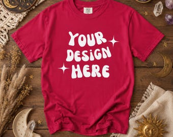 以下が含まれることがあります： 白い文字で「YOUR DESIGN HERE」と書かれた赤いTシャツ。木製の表面に、太陽、月、クリスタルなどの天体モチーフの装飾が施されています。カスタムデザイン用の無地のTシャツです。