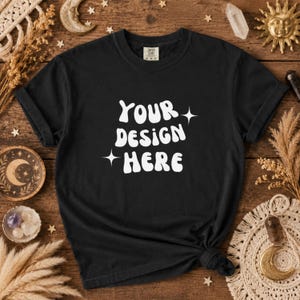 Puede incluir: Camiseta negra con el texto blanco "YOUR DESIGN HERE" en una fuente estilizada. La camiseta está anudada en la parte inferior y tiene mangas cortas. Se muestra sobre una superficie de madera con elementos decorativos celestiales.