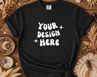 以下が含まれることがあります： 白い「YOUR DESIGN HERE」の文字が特徴的な黒いTシャツ。Tシャツは裾で結ばれており、半袖です。木製の表面に、装飾的な天体の要素とともに表示されています。