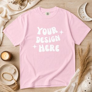 Puede incluir: Camiseta rosa claro con el texto blanco "YOUR DESIGN HERE". La camiseta de cuello redondo tiene mangas cortas. La camiseta se exhibe sobre una superficie de madera, rodeada de elementos decorativos.