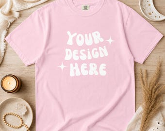 以下が含まれることがあります： 白の「YOUR DESIGN HERE」の文字が入った、明るいピンク色のTシャツ。クルーネックのTシャツは半袖です。木製の表面に飾られており、キャンドルや三日月など、装飾品に囲まれています。