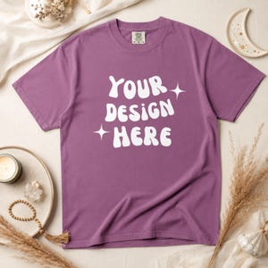 Puede incluir: Una camiseta morada con el texto blanco "YOUR DESIGN HERE" en una fuente estilizada, adornada con estrellas. La camiseta tiene cuello redondo y se muestra sobre una superficie neutra. La imagen incluye elementos decorativos como una vela y flores secas.