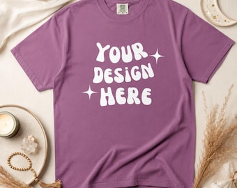 以下が含まれることがあります： 白い「YOUR DESIGN HERE」の文字がスタイリッシュなフォントでプリントされた紫色のTシャツ。Tシャツはクルーネックで、星のアクセントが付いています。ニュートラルな表面に表示され、キャンドルやドライフラワーなどの装飾要素が含まれています。