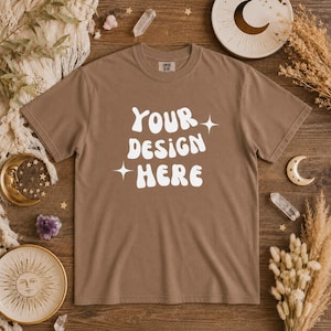 Puede incluir: Una camiseta marrón con el texto "YOUR DESIGN HERE" en blanco, con detalles de estrellas. La camiseta es de la marca Comfort Colors. La imagen es una maqueta de producto, probablemente para ropa personalizada.