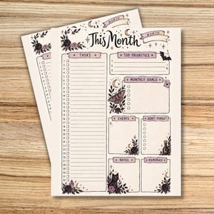 Spooky-cute printable monthly planner pages displayed on a wooden background, featuring black cats, bats, roses, moons, stars, crystals, and a moth. The main page includes sections for tasks, top priorities, monthly goals, events, reminders, notes, and memories.