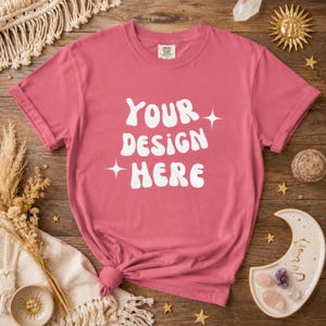 Puede incluir: Una camiseta de color rosa polvoriento con las mangas enrolladas. La camiseta presenta el texto "YOUR DESIGN HERE" en una fuente retro blanca. La camiseta está anudada en la parte inferior. La camiseta está sobre una superficie de madera.
