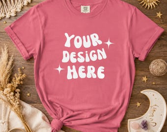 以下が含まれることがあります： 袖をまくったダスティローズ色のTシャツ。Tシャツには、白いレトロフォントで「YOUR DESIGN HERE」という文字がプリントされています。Tシャツは裾で結ばれています。Tシャツは木製の表面にあります。