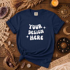 Puede incluir: Camiseta azul marino con el texto blanco "YOUR DESIGN HERE" y acentos de estrellas. La camiseta está anudada en la parte inferior. La imagen muestra la camiseta sobre una superficie de madera, rodeada de objetos decorativos.