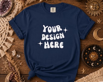 以下が含まれることがあります： 白文字で「YOUR DESIGN HERE」と星のアクセントが入ったネイビーブルーのTシャツ。Tシャツは裾で結ばれています。画像は、木製の表面にTシャツが置かれ、装飾品に囲まれている様子を示しています。