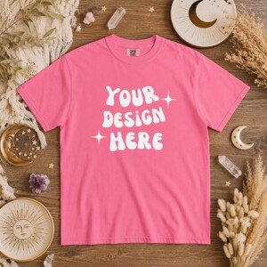 Puede incluir: Una camiseta rosa vibrante con el texto blanco "YOUR DESIGN HERE" en letras estilizadas. La camiseta se muestra sobre una superficie de madera, rodeada de elementos decorativos que incluyen cristales y decoración de temática celestial.
