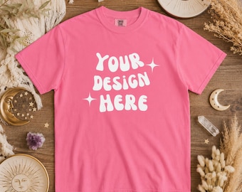 以下が含まれることがあります： 白文字で「YOUR DESIGN HERE」と書かれた、鮮やかなピンク色のTシャツ。木製の表面に置かれ、クリスタルや天体モチーフの装飾など、装飾的な要素に囲まれています。