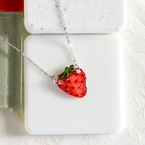 May include: A silver chain necklace with a red and green strawberry pendant. The strawberry is painted with white seeds and a green stem.