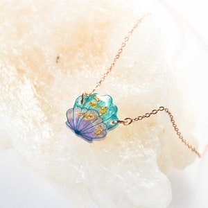 May include: A delicate gold chain necklace with a seashell pendant. The seashell is made of resin and features a blue and purple color gradient with gold flecks.