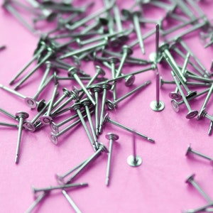 Könnte beinhalten: Nahaufnahme einer großen Menge silberfarbener Stecknadeln, die auf einer rosa Oberfläche verstreut sind.