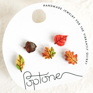 Könnte beinhalten: Satz mit fünf Ohrringen zum Thema Herbst, bestehend aus einer braunen Eichel und vier Blättern in Rot-, Orange-, Gelb- und Grüntönen. Die Ohrringe werden auf einer weißen Karte mit dem Text "Handgefertigter Schmuck für die Vibrant Quirky" und dem Wort "Optome" in Kursivschrift präsentiert.