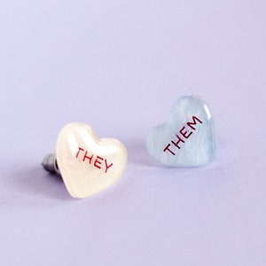 Könnte beinhalten: Zwei herzförmige Pins mit den Worten "THEY" und "THEM" in Rot auf einem hellvioletten Hintergrund.