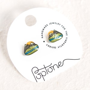 Puede incluir: Un par de pendientes con temática de montaña con un sol amarillo, montañas verdes y un lago azul. Los pendientes están en una tarjeta blanca con el texto "Joyería hecha a mano para los vibrantemente extravagantes Poptone".