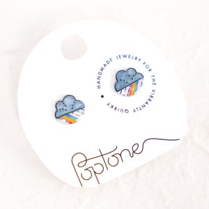 May include: A pair of handmade ceramic earrings featuring a blue cloud with a rainbow underneath. The earrings are on a white card with the text "Handmade Jewelry for the Vibrantly Quirky" in a circle.