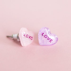 May include: Two pink heart-shaped earrings with silver posts. One earring says "XOXO" and the other says "LOVE".