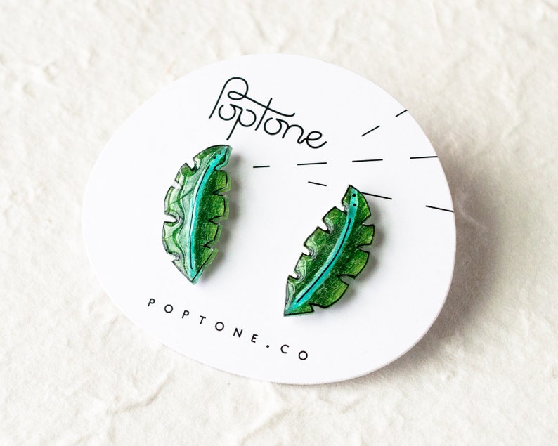 May include: A pair of green and blue leaf-shaped earrings with a white background. The earrings are made of resin and have a glossy finish. The text "Poptone.co" is printed on the background.