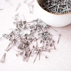 May include: A large quantity of silver-toned metal tacks or brads scattered on a white surface. The tacks have a flat head and a pointed end.