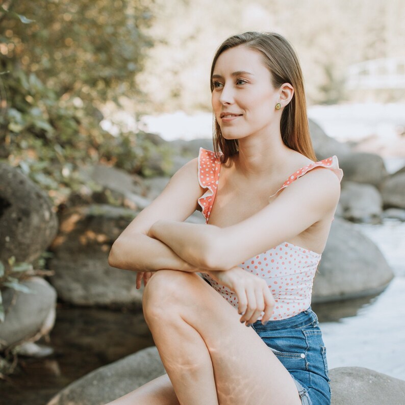 Puede incluir: Una mujer con un traje de ba&ntilde;o de lunares rosa y blanco con una falda vaquera azul, sentada en una roca junto a un arroyo.