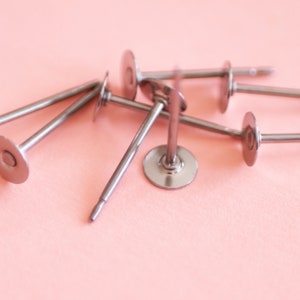 May include: A close-up of six silver metal earring posts with round, flat backs. The posts are arranged in a pile on a pink surface.