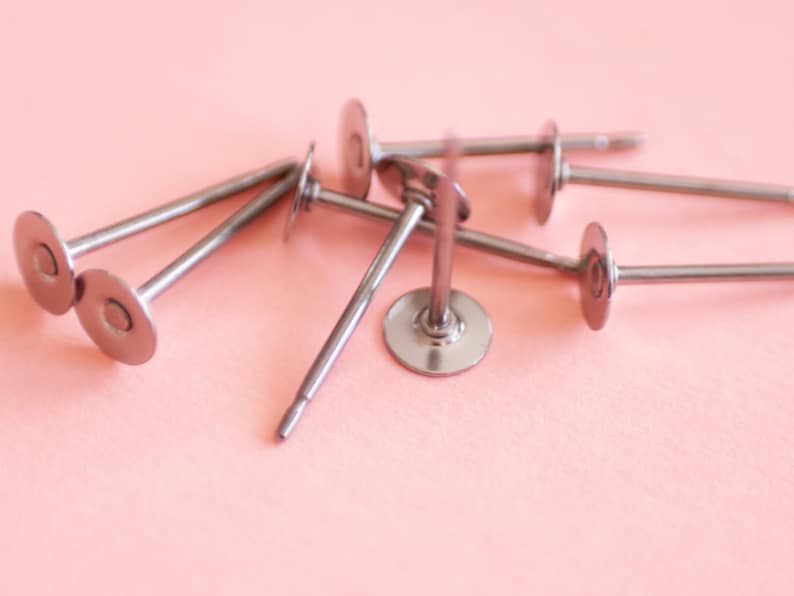 May include: A close-up of six silver metal earring posts with round, flat backs. The posts are arranged in a pile on a pink surface.