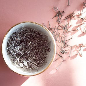 May include: A white ceramic bowl filled with silver tacks or upholstery tacks. The bowl is on a pink surface and there are more tacks scattered around the bowl.