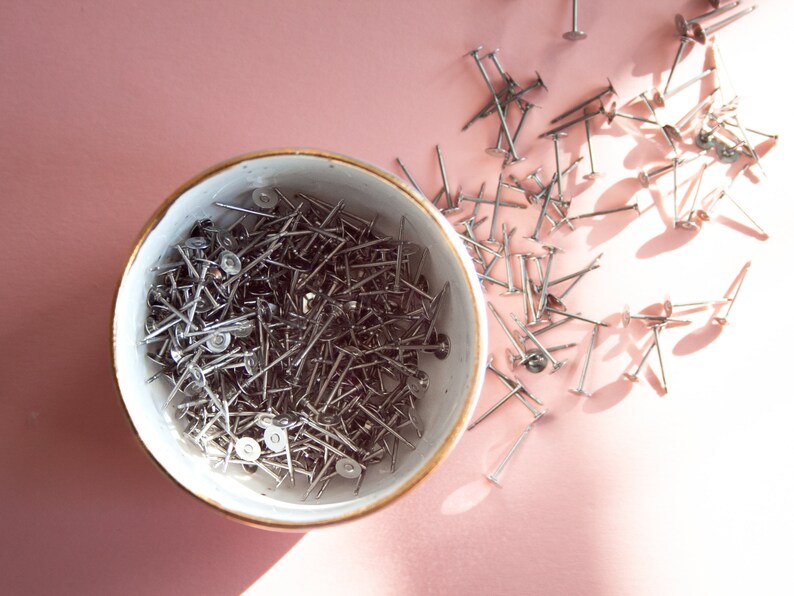 May include: A white ceramic bowl filled with silver tacks or upholstery tacks. The bowl is on a pink surface and there are more tacks scattered around the bowl.