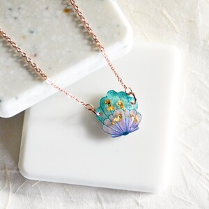 May include: A delicate rose gold chain necklace with a teal, purple, and gold seashell pendant. The seashell has a layered, iridescent look with flecks of gold.