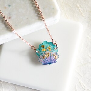 May include: A delicate rose gold chain necklace with a seashell pendant. The seashell is made of resin and features a blue and purple gradient with gold flecks.