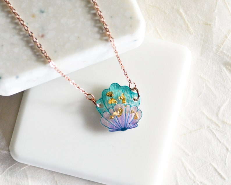 May include: A delicate rose gold chain necklace with a seashell pendant. The seashell is made of resin and features a blue and purple gradient with gold flecks.