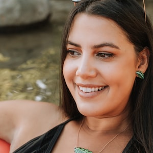 May include: A woman with long dark hair wearing a black swimsuit with a green leaf pendant necklace and matching earrings. She is smiling and looking to the side.
