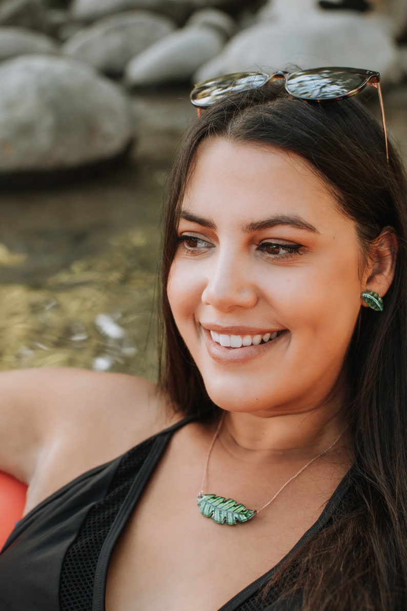 May include: A woman with long dark hair wearing a black swimsuit with a green leaf pendant necklace and matching earrings. She is smiling and looking to the side.