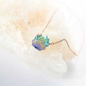 May include: A delicate gold chain necklace with a seashell pendant. The seashell is made of resin and features a blue and purple iridescent finish with gold flecks.