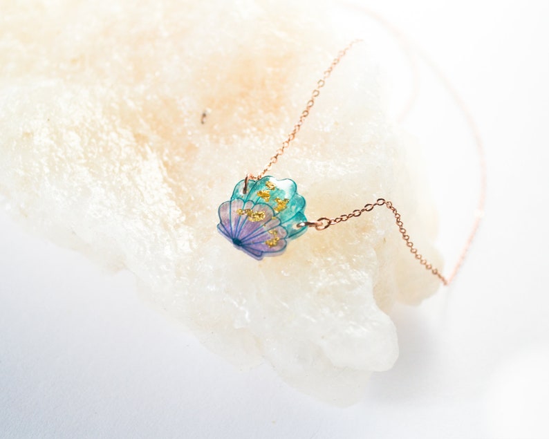 May include: A delicate gold chain necklace with a seashell pendant. The seashell is made of resin and features a blue and purple iridescent finish with gold flecks.